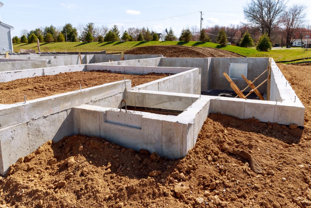Fondation en béton d'une nouvelle maison en construction. Des ancrages métalliques dépassent des murs, la terre est retournée, ciel bleu.