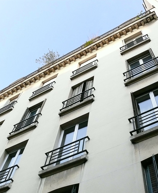 Façade d'immeuble parisien haussmannien beige, fenêtres à balcons en fer forgé noir, et végétation sur la corniche sous ciel clair.