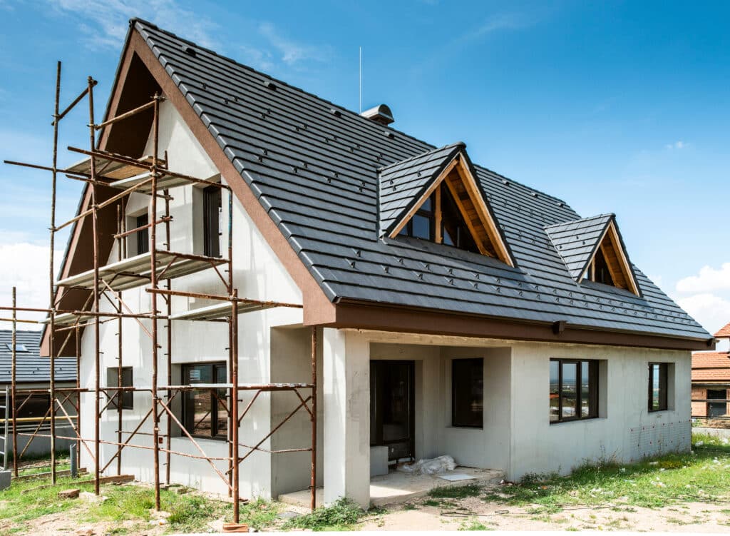 Maison neuve en construction avec échafaudage rouillé, toiture en tuiles grises foncées, et murs crépis blancs sous un ciel bleu.