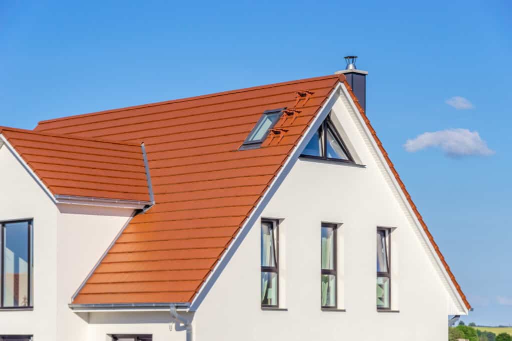 Toit de maison moderne en tuiles rouges, façade blanche, cheminée noire et fenêtres sous ciel bleu clair avec nuages.