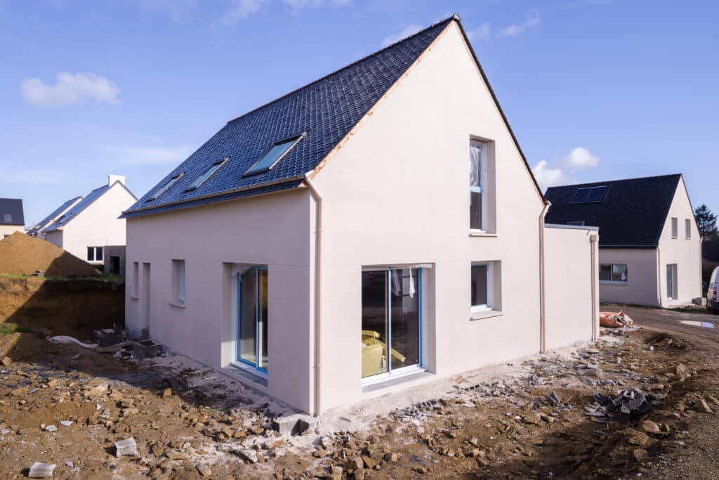 Nouvelle maison en construction avec façade beige, toit sombre en tuiles et fenêtres de toit. Le terrain est non aménagé, avec terre et gravats.