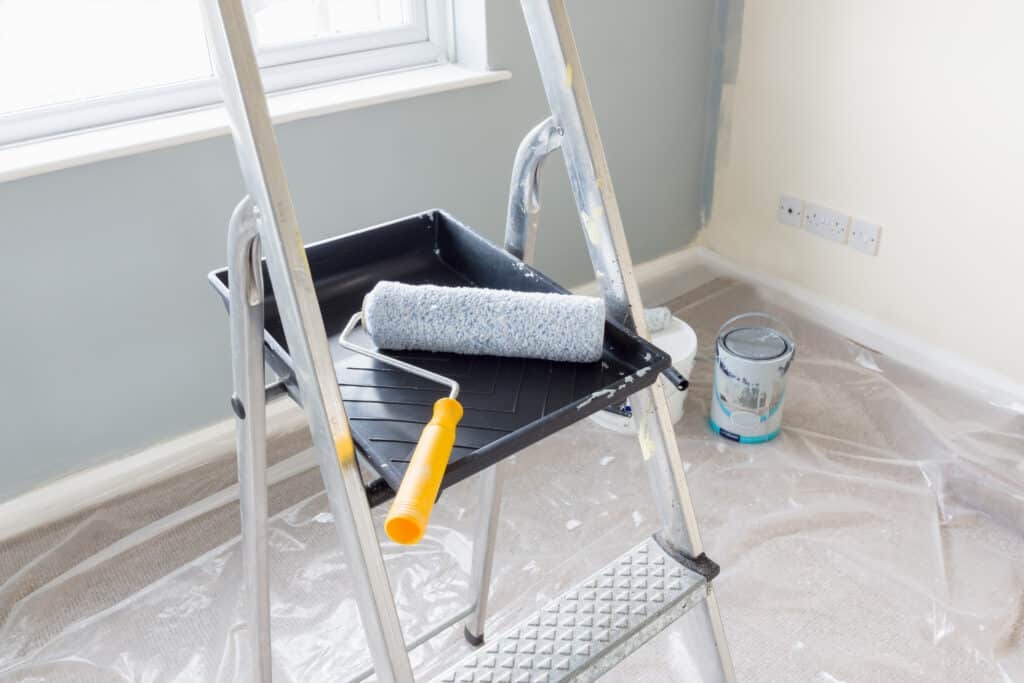 Échelle, rouleau et bac de peinture dans pièce en rénovation. Murs gris partiels, sol protégé par bâches, pots de peinture au sol.