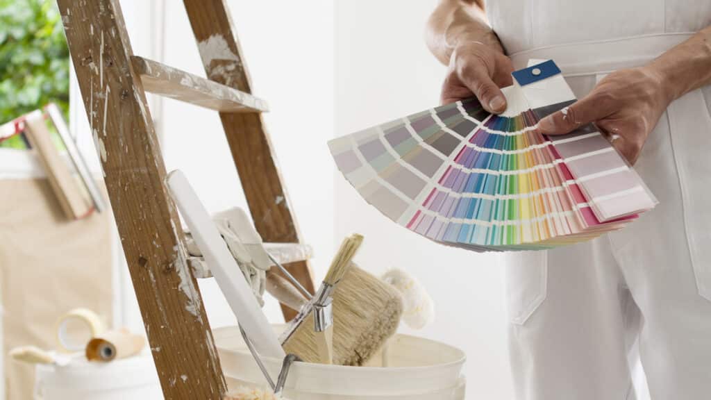Peintre en uniforme blanc tenant un nuancier pour choisir les couleurs. Une échelle éclaboussée et des outils de peinture sont à ses côtés.