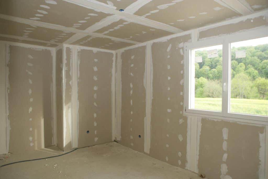 Pièce de maison en construction avec murs et plafond en placo beige enduits de blanc. Grande fenêtre PVC blanche donnant sur un paysage verdoyant.