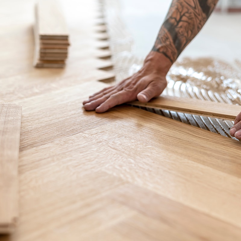 Mains posant un parquet clair en chevrons sur une couche d'adhésif. Des lattes de bois sont empilées à proximité.