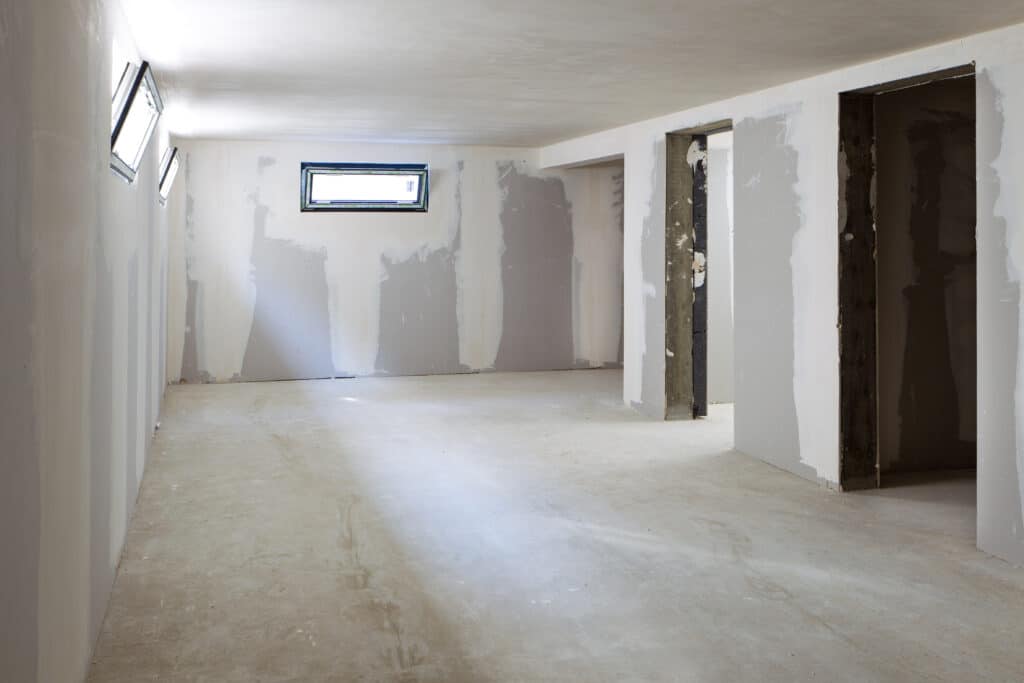 Pièce en rénovation avec murs en placo partiellement enduits et sol en béton brut. Plusieurs fenêtres et ouvertures de portes inachevées.