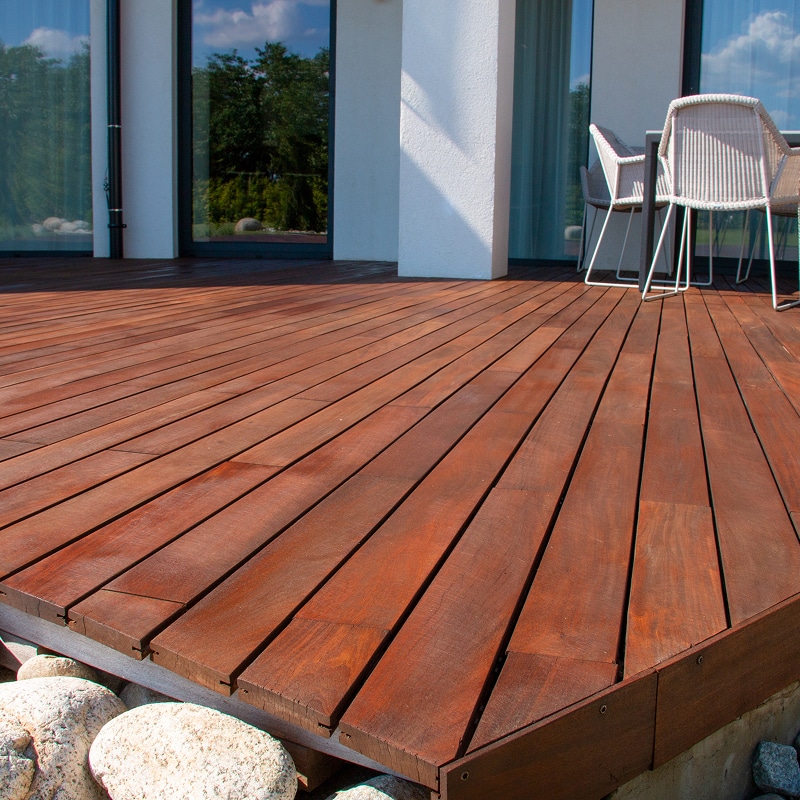 Grande terrasse en bois brun-rouge avec des planches larges, devant une maison moderne aux baies vitrées. Des pierres claires bordent le deck.