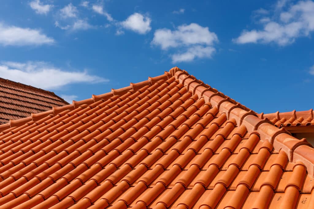 Toit traditionnel en tuiles de terre cuite rouge-orangé sous un ciel bleu clair parsemé de nuages blancs. Deux sections de toit visibles.